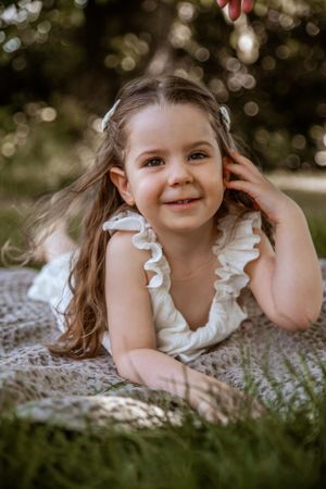 Young child in white ruffled dress lying on blanket outdoors with bokeh background and natural lighting.