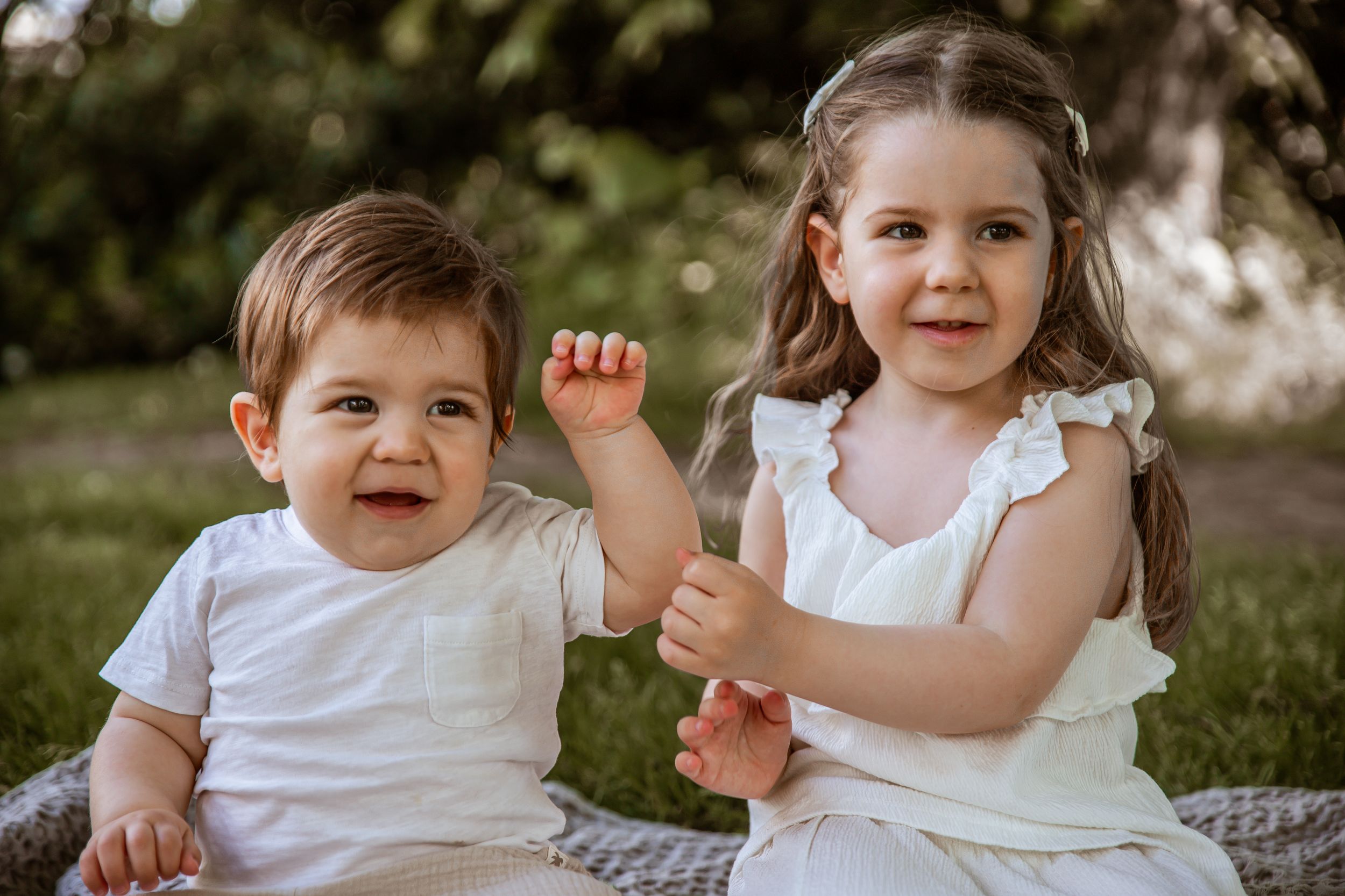 Two children in white clothing sit together outdoors on a blanket during a sunny day.