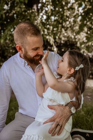 Sweet father-daughter moment captured outdoors with joyful expressions and natural lighting under trees.