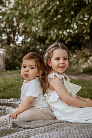 Two children in white outfits sit back-to-back on blanket in garden setting with soft natural lighting.