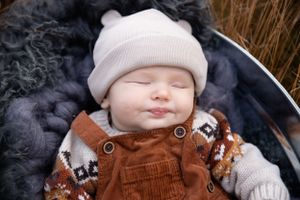 Small baby wearing a white beanie and brown corduroy overalls sleeping peacefully on dark furry blanket in winter.