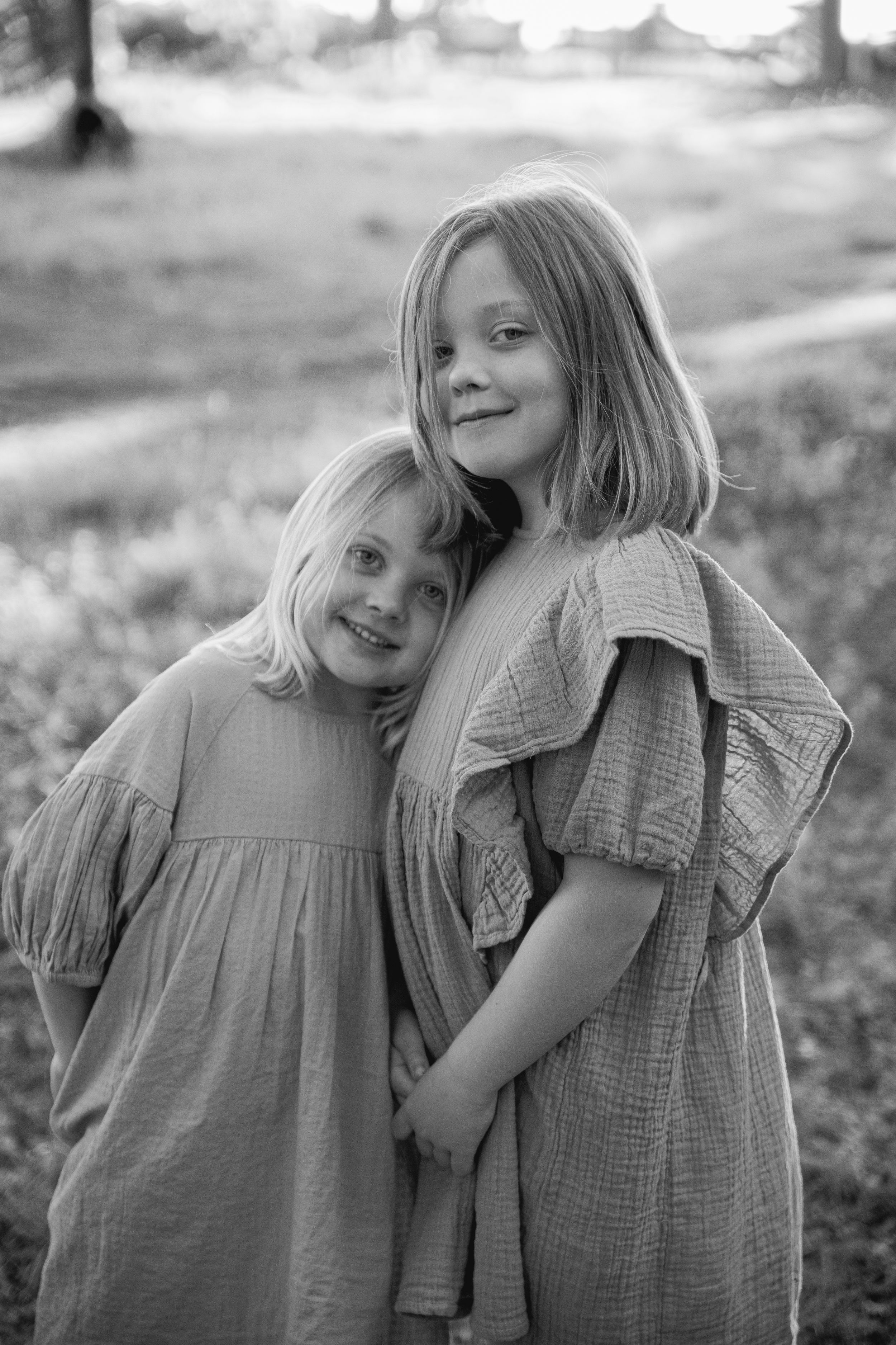 Black and white portrait of two young children in loose cotton dresses hugging and smiling outdoors.