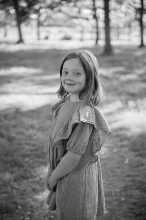 A black and white portrait of a young child in a ruffled dress standing outdoors among trees with soft bokeh background.