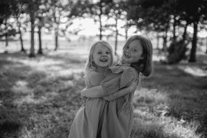 Two young children in matching dresses embrace and laugh together in a sunlit wooded area, captured in black and white.