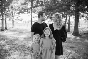 A family of four poses together outdoors in black and white, standing amongst trees in a natural setting.