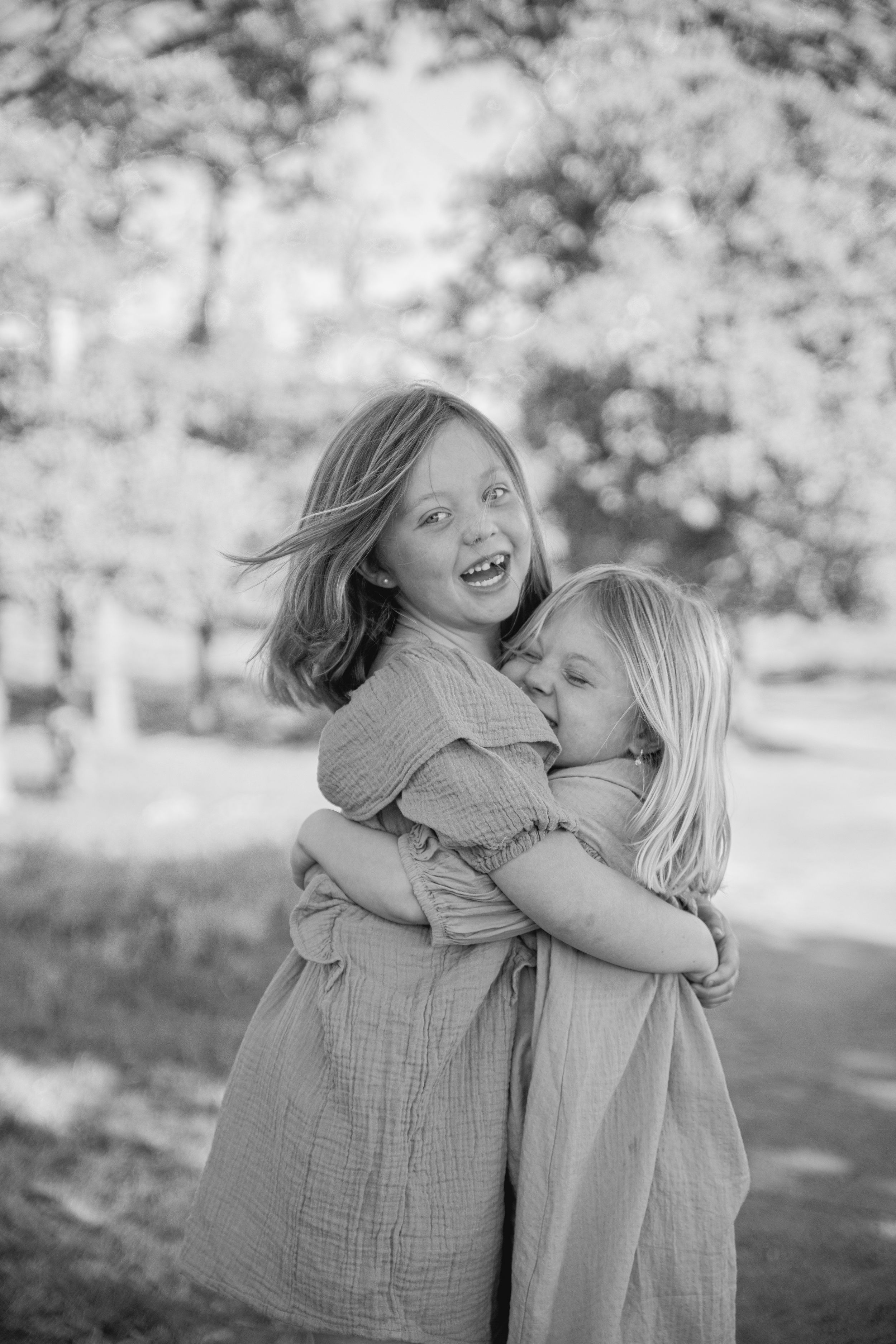Two kids in casual dresses share a joyful embrace while playing outdoors in black and white photography.