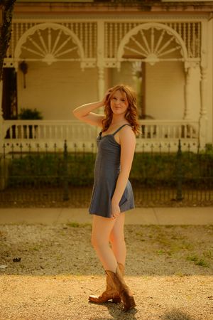 A person in a gray sundress and brown boots poses in front of a Victorian-style porch with ornate white railings at sunset.