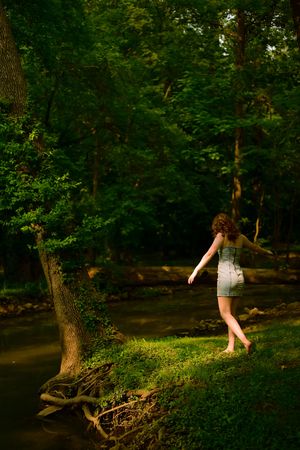 A person in a dress balances on a tree root by a creek in a lush green forest setting on a summer day.