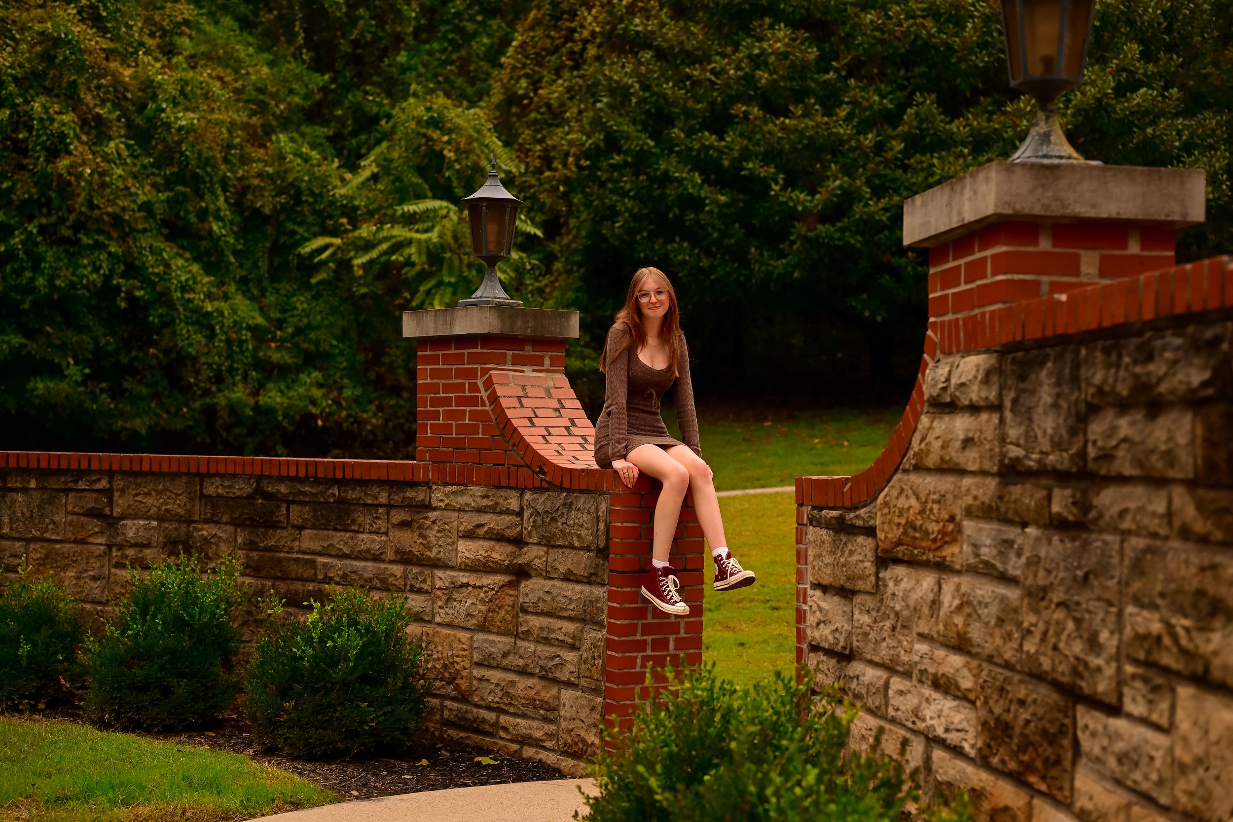A person sits on a decorative brick and stone wall at dusk in a garden setting with lush greenery in the background.
