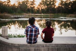 Two people sitting together on a concrete wall overlooking a tranquil lake at sunset with trees in the background.
