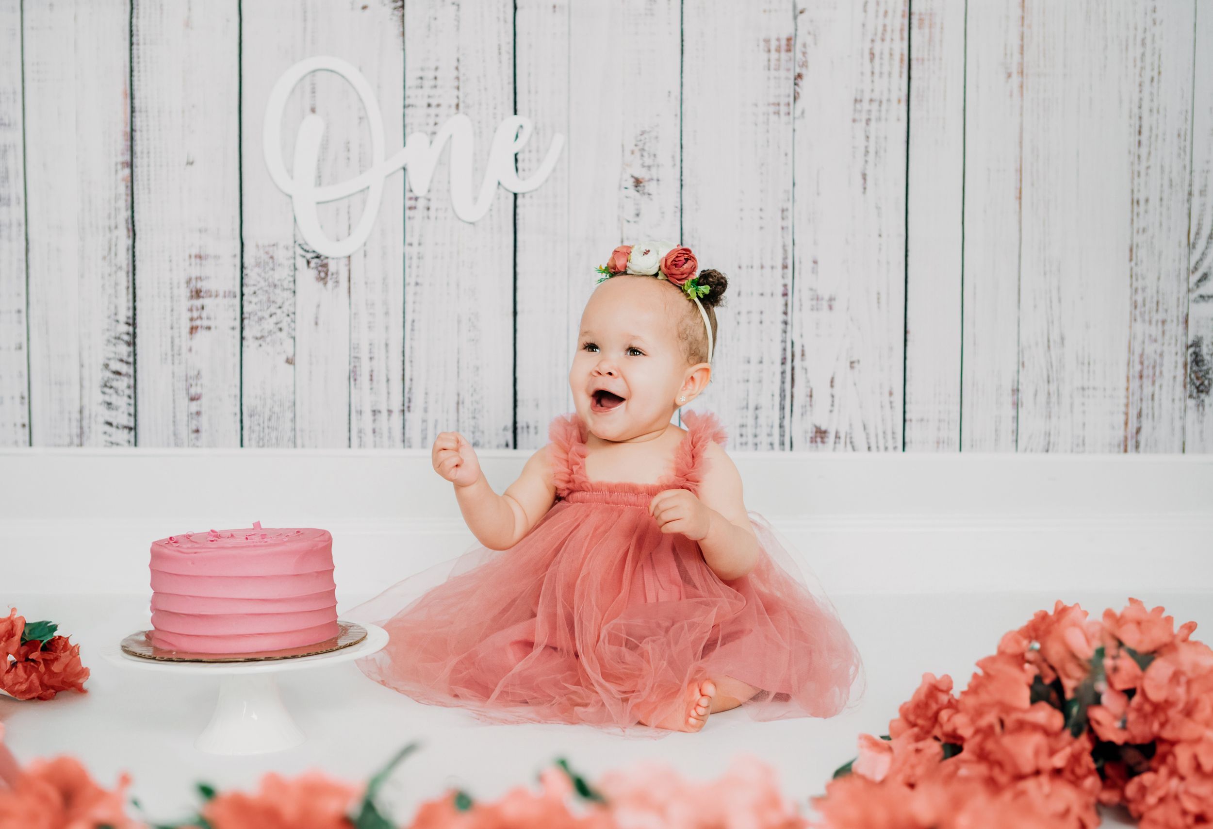 A small figure in a coral tulle dress sits beside a pink birthday cake surrounded by matching coral flowers against a white backdrop.