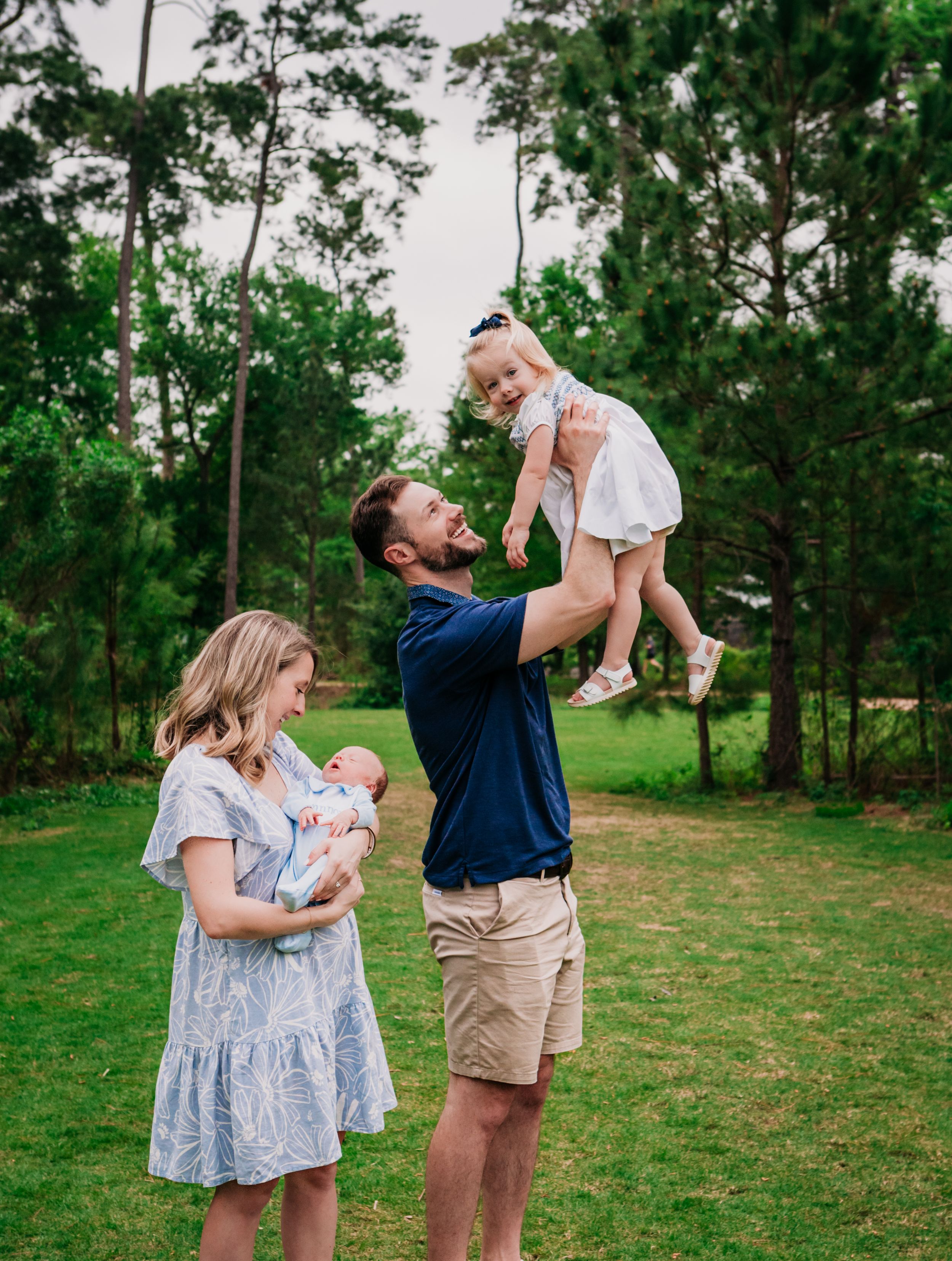 Family enjoying a playful outdoor moment together on a green lawn surrounded by tall pine trees.