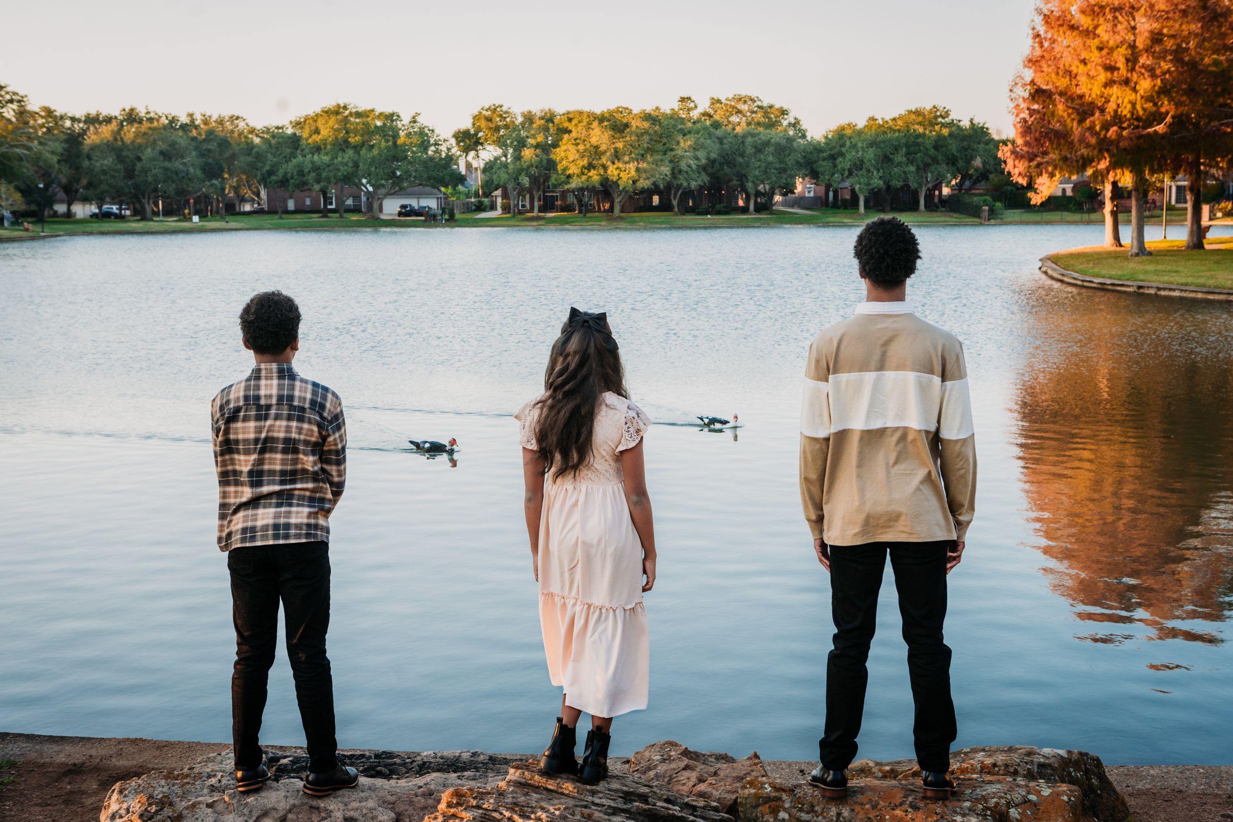 Three figures stand at the edge of a tranquil lake during autumn with trees reflecting golden colors in the water.