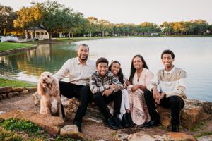 A group sits together with their goldendoodle dog by a serene lake at sunset in a park setting.