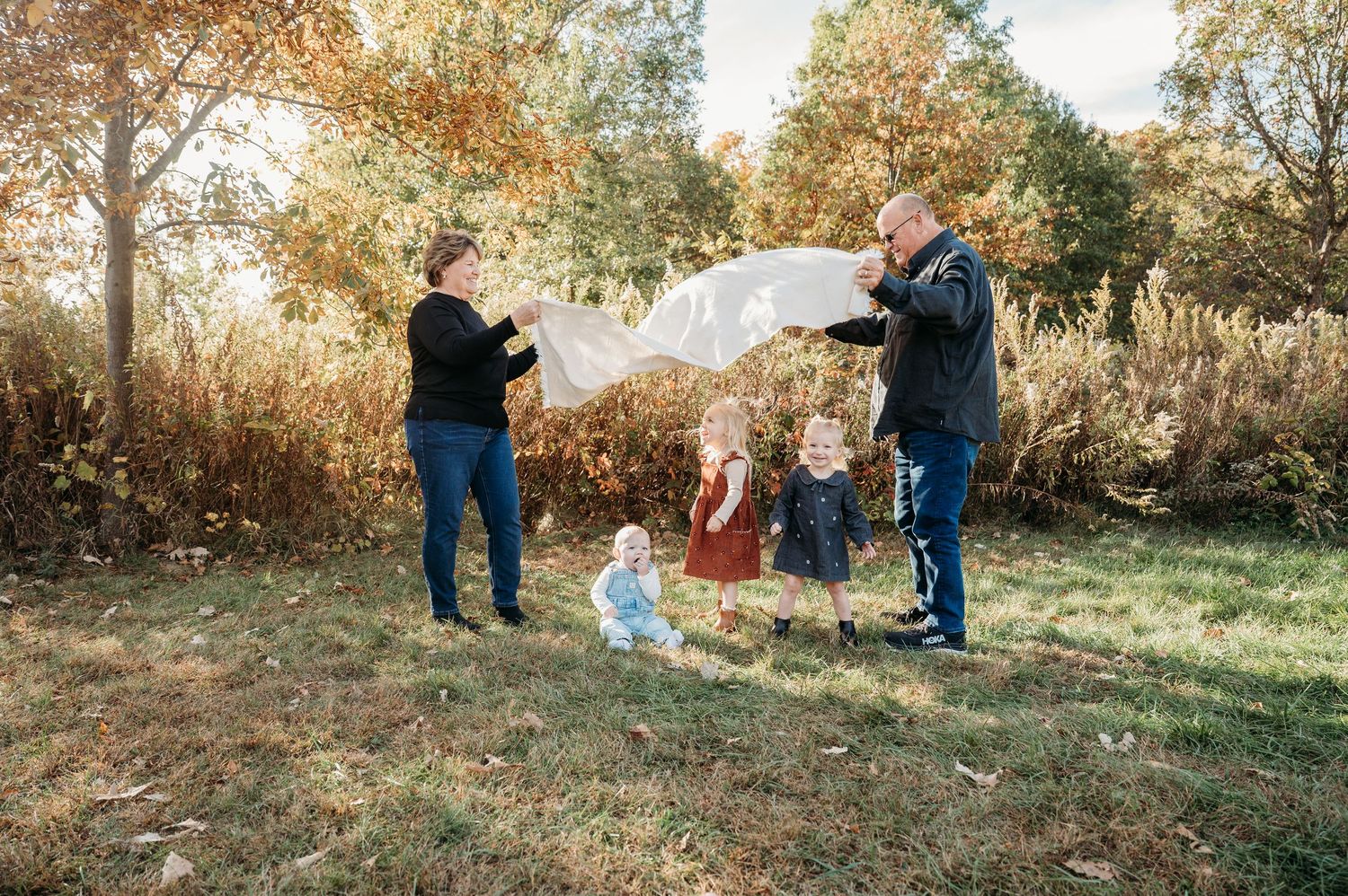 Fall photo with the sun peaking through. Two grandparents waving a blanket with their infant grandkids playing underneath it.