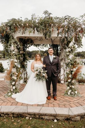 A wedding couple stands under an ornate floral arch with cascading greenery and white flowers.