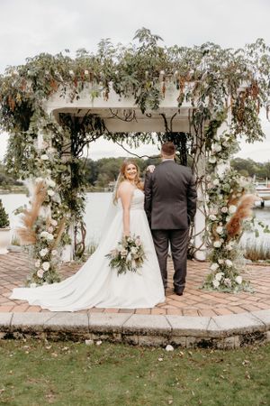 A wedding couple stands under an elegant floral arch decorated with greenery and white flowers.