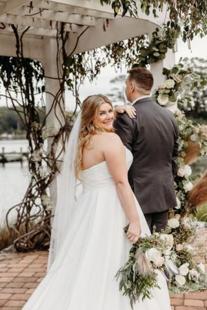 Couple in wedding attire standing under decorated pergola with greenery.
