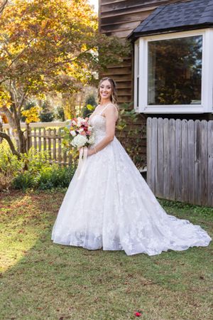 Bride in elegant lace ball gown poses by rustic building with fall foliage.
