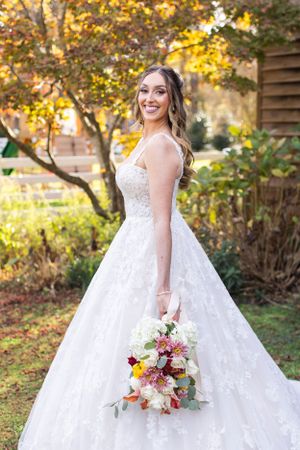 Bride holds colorful bouquet while posing in white ball gown against autumn backdrop.
