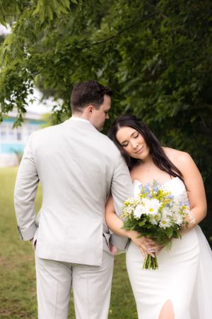 Romantic wedding portrait under lush green trees with bride holding floral bouquet.
