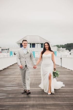 Bride and groom in light colored attire walking along seaside boardwalk.