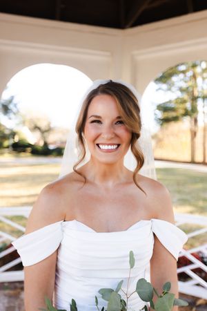 Bride in white off-shoulder dress smiles radiantly while holding eucalyptus bouquet during outdoor wedding photoshoot.