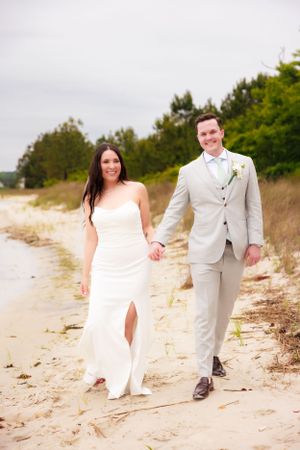 Newlyweds walking hand in hand along sandy beach in light colored wedding attire.