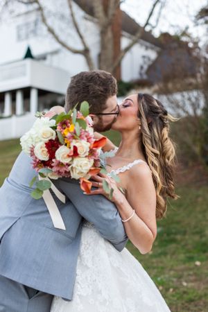 A romantic wedding kiss with the couple in grey suit and white dress holding a floral bouquet.