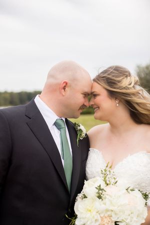 Bride and groom touching foreheads and smiling, bride holding white bouquet, groom in black suit with green tie.