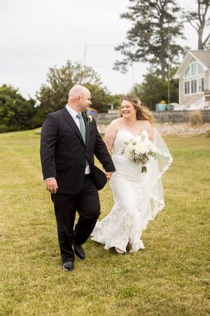 Bride and groom walk hand-in-hand on green lawn, she in lace gown with bouquet, he in black suit with green tie.