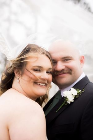 Smiling bride and groom posing closely together outdoors, bride wearing veil, groom in black tuxedo with white boutonniere.