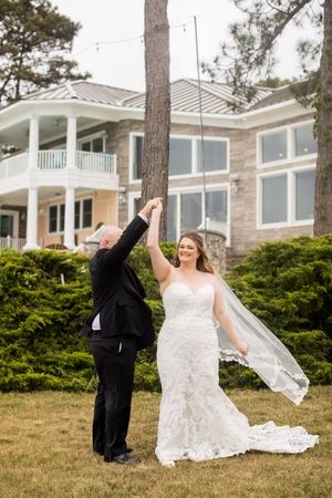 Bride and groom dancing outdoors on lawn in front of elegant home, bride in lace gown with veil flowing.