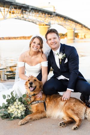 Wedding couple poses with their happy golden retriever dog against a scenic bridge backdrop at sunset.