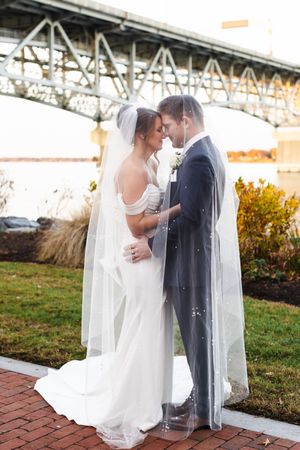 Romantic wedding couple embraces on brick pathway under autumn sky with flowing white veil.