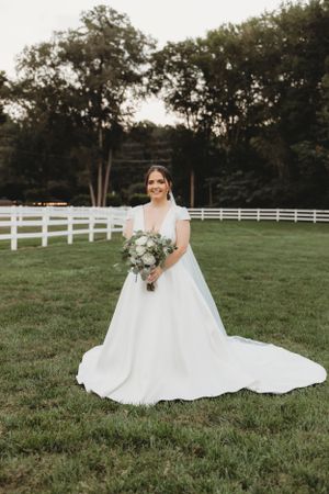 A bride in a classic white gown holds a bouquet on a scenic farm property.
