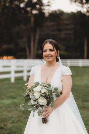 A radiant bride poses with her bouquet against white fence and trees at dusk.