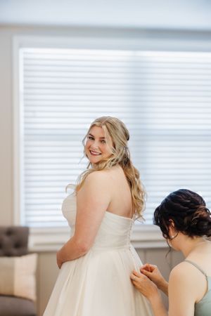 A bride in a strapless wedding gown smiles while getting dressed for her wedding day.