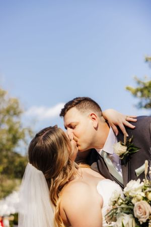 A romantic wedding kiss captured outdoors against a blue sky with delicate floral details.