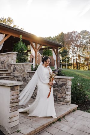 Bride in elegant white dress and flowing cape veil stands on stone patio steps during golden sunset hour.
