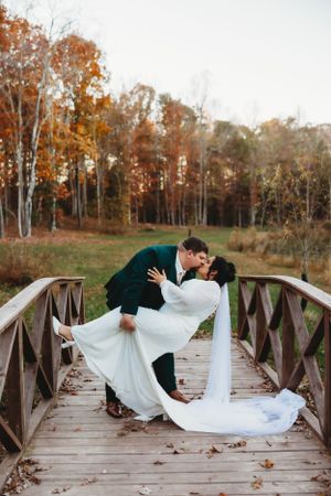 A romantic couple in wedding attire share a dramatic dip kiss on a wooden bridge surrounded by autumn foliage.