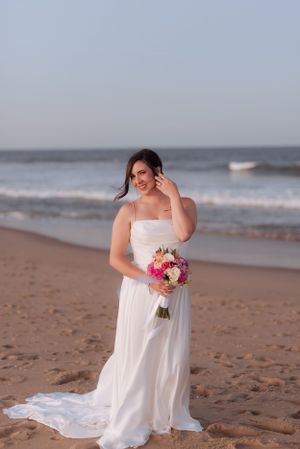 A bride in a flowing white dress holds a vibrant pink and white bouquet while standing on a beach at sunset.