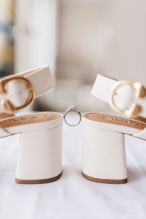 Elegant engagement ring balanced between white block heeled shoes on light fabric background.