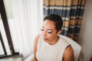 Bride with short hair looks down thoughtfully near window during wedding preparations.