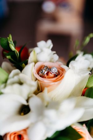 Wedding rings nestled in pale peach rose surrounded by white flowers in bridal bouquet.