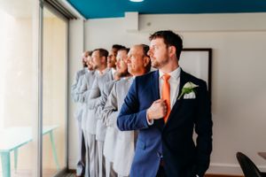 Groomsmen in navy and gray suits stand by window in wedding preparation room.