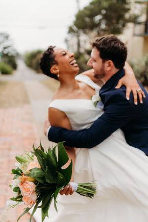 Wedding couple shares a laugh while embracing, with peach and white bouquet in hand.