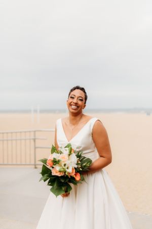 A joyful bride in a classic white wedding dress holds a peach and white bouquet against an overcast sky.