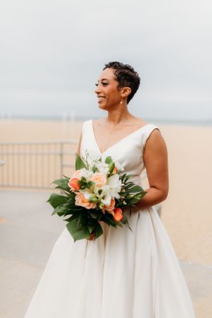 Bride in sleeveless white gown smiles while holding tropical bouquet with peach and white flowers.