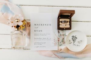 Wedding details including invitation, ring box, and floral accents arranged on white wooden surface.